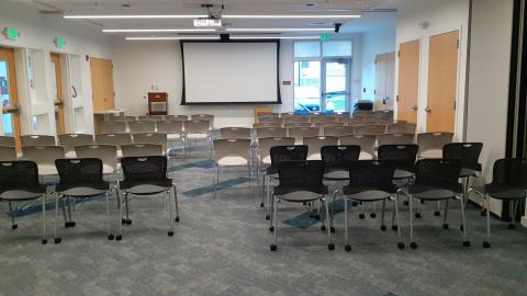 Community Room fully opened, viewed from the back wall with chairs set up facing the projection screen. 