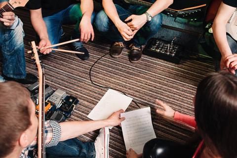 image of several people sitting in a circle, each holding a different musical instrument. Two people in the foreground are reading sheet music.