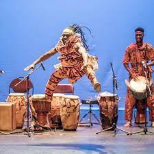 A dancer in traditional Guinean dress jumps in the air as a drummer, also in traditional dress, plays behind him.