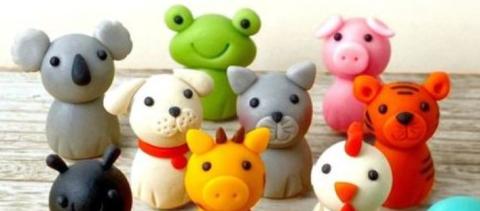 Photo of a dozen little creatures made in a similarly cartoonish style, out of air-dried clay of many colors. They are arranged in a casual group on a wooden table, set against a white wall, all facing the viewer.