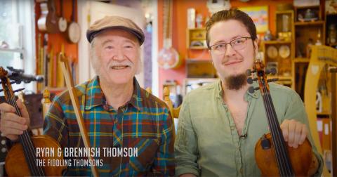 A wide frame, color portrait of Ryan and Brennish Thomson, aka "The Fiddling Thomsons," in which they smile at the camera and sit holding their instruments. Their names are in the lower left corner of the image in light grey text.