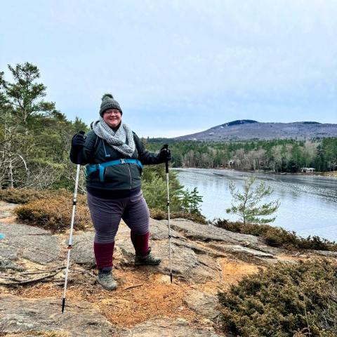 Paige Emerson stands with hiking poles near a pond with a mountain in the background