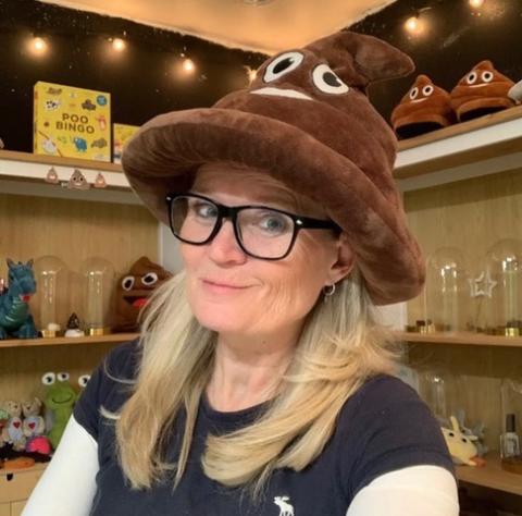 Photo of Susie Maguire of The Poop Museum, smiling at the camera with a trademark Poop Emoji hate on her head. Behind her are glass jars filled with assorted items.