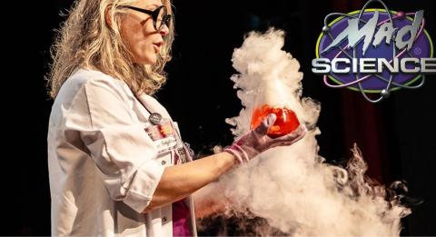 A photo of a scientist in a lab coat, gloves, and thick black-rimmed glasses, holding up a beaker with bright orange liquid inside, as it spews out thick white vapor. In the upper right corner of the image is the logo for Mad Science of Maine.