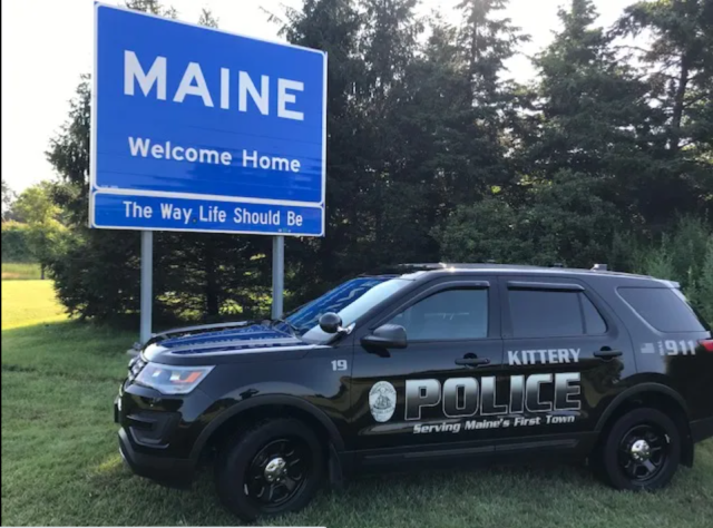 Navy blue Kittery Police cruiser SUV parked in front of the blue 'Welcome to Maine' sign on I-95. 