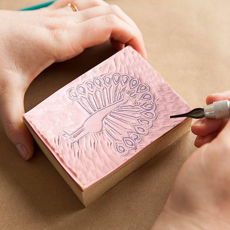 Two hands carve a peacock design out of the face of a rubber stamp.