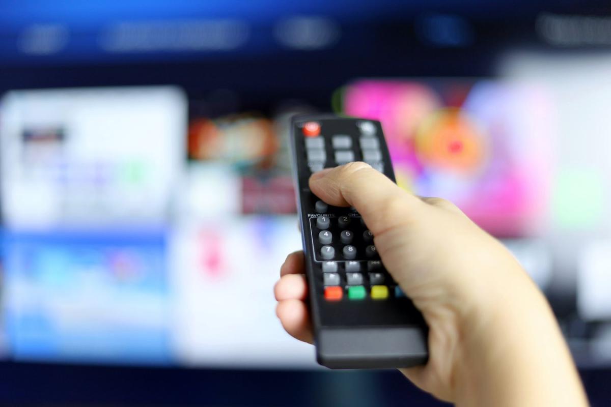 A hand holds a remote control in front of an out-of-focus smart TV with several streaming apps on the screen.