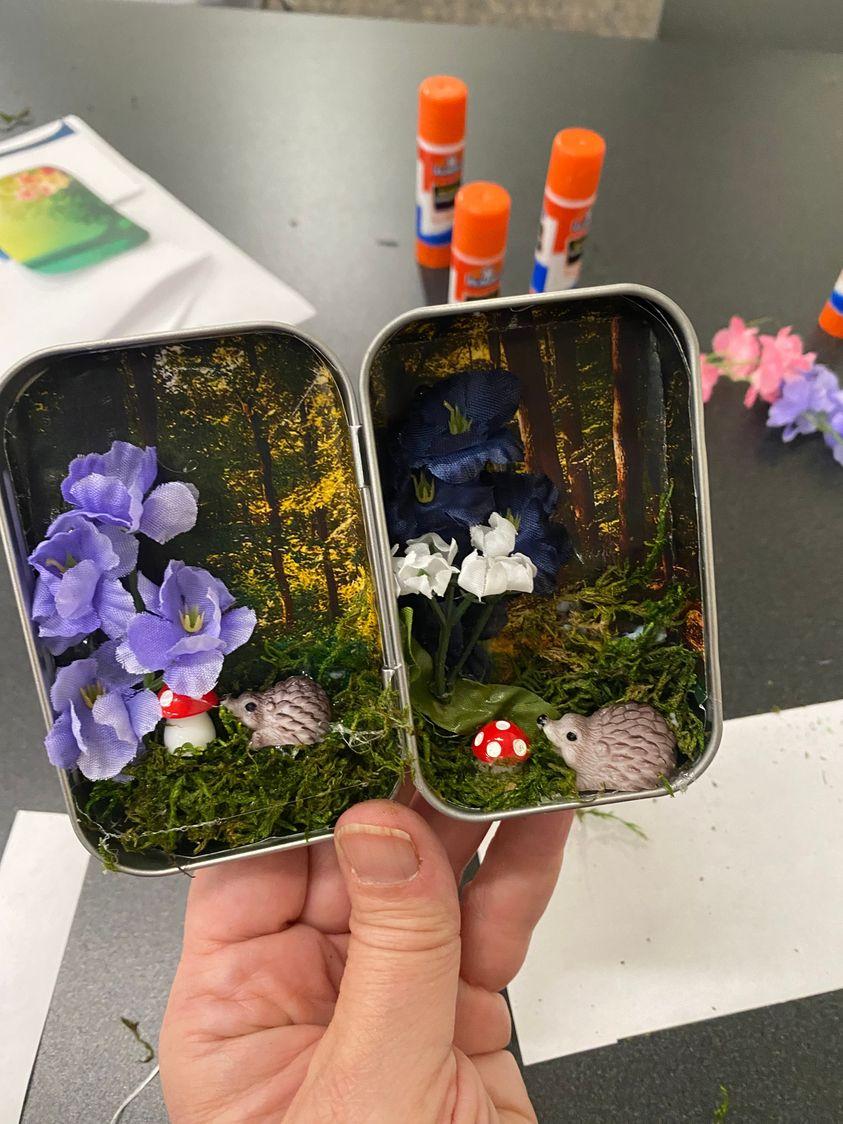 A small metal tin with a garden scene inside, with mushrooms, trees, and other art. Held in a light-skinned hand over a table with supplies like glue sticks on it.