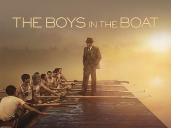 A man in a suit and hat stands on a dock with an 8-person crew boat in the water.