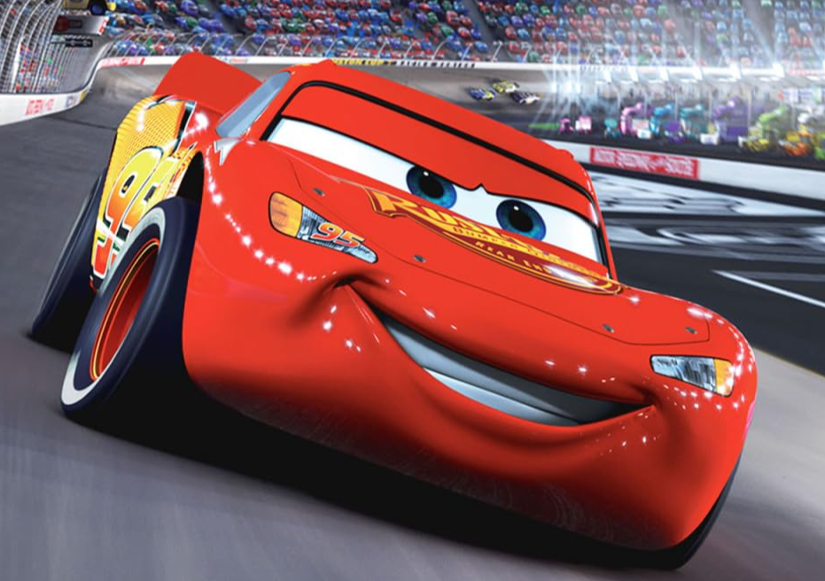 A closeup action freezeframe from the movie 'Cars,' showing the main character, Lightning McQueen, racing around the track toward the viewer. He nearly fills the frame, with a dozen other cars visible far behind him on the last turn. He grins like he knows he's going to win the race.