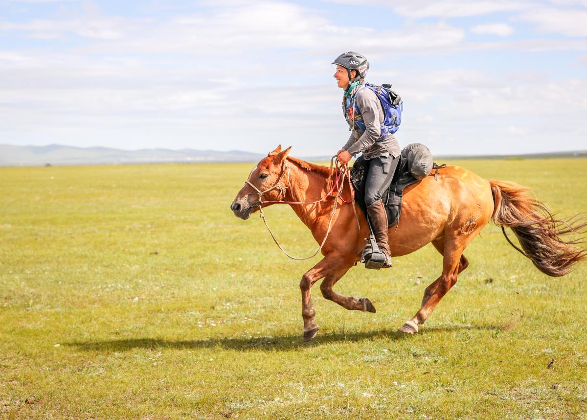 A photo of a rider on a horse, galloping from the right to the left across the image, on a wide grassy plain under a blue and lightly cloudy sky.